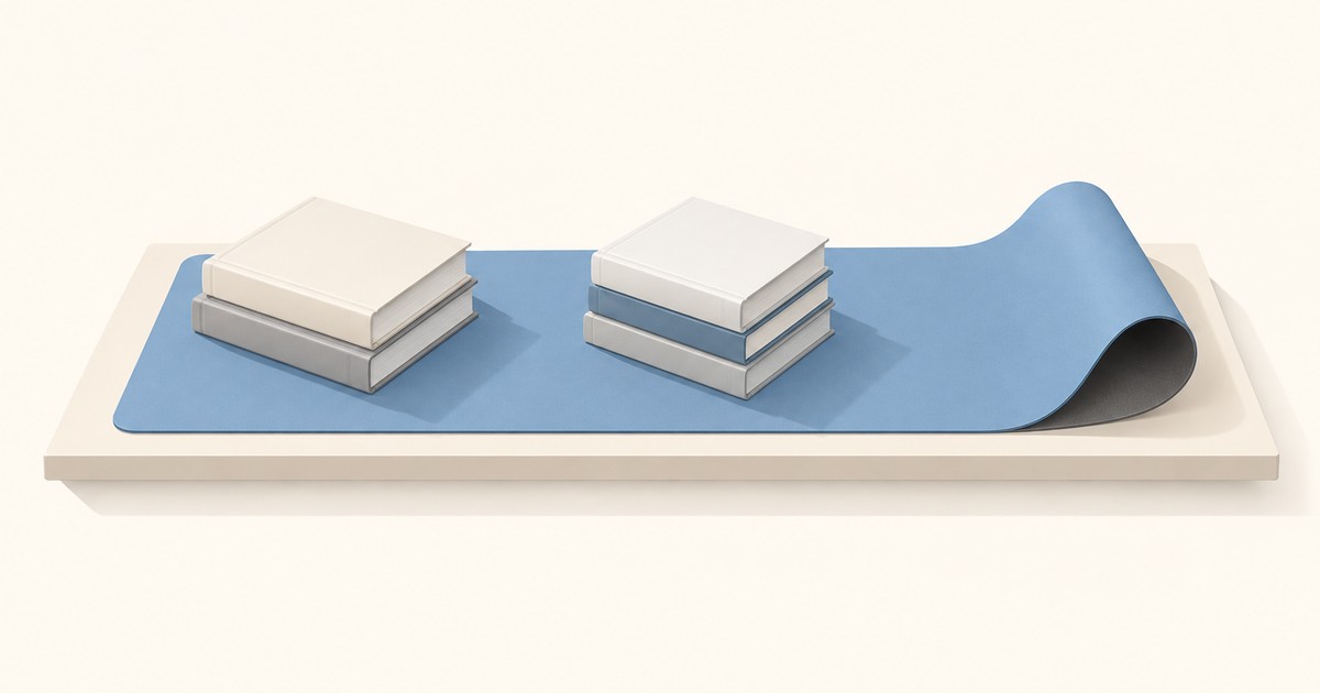 Blue desk mat on a cream desk weighed down by two stacks of plain hardcover books while its right end still curls upward with a gray underside